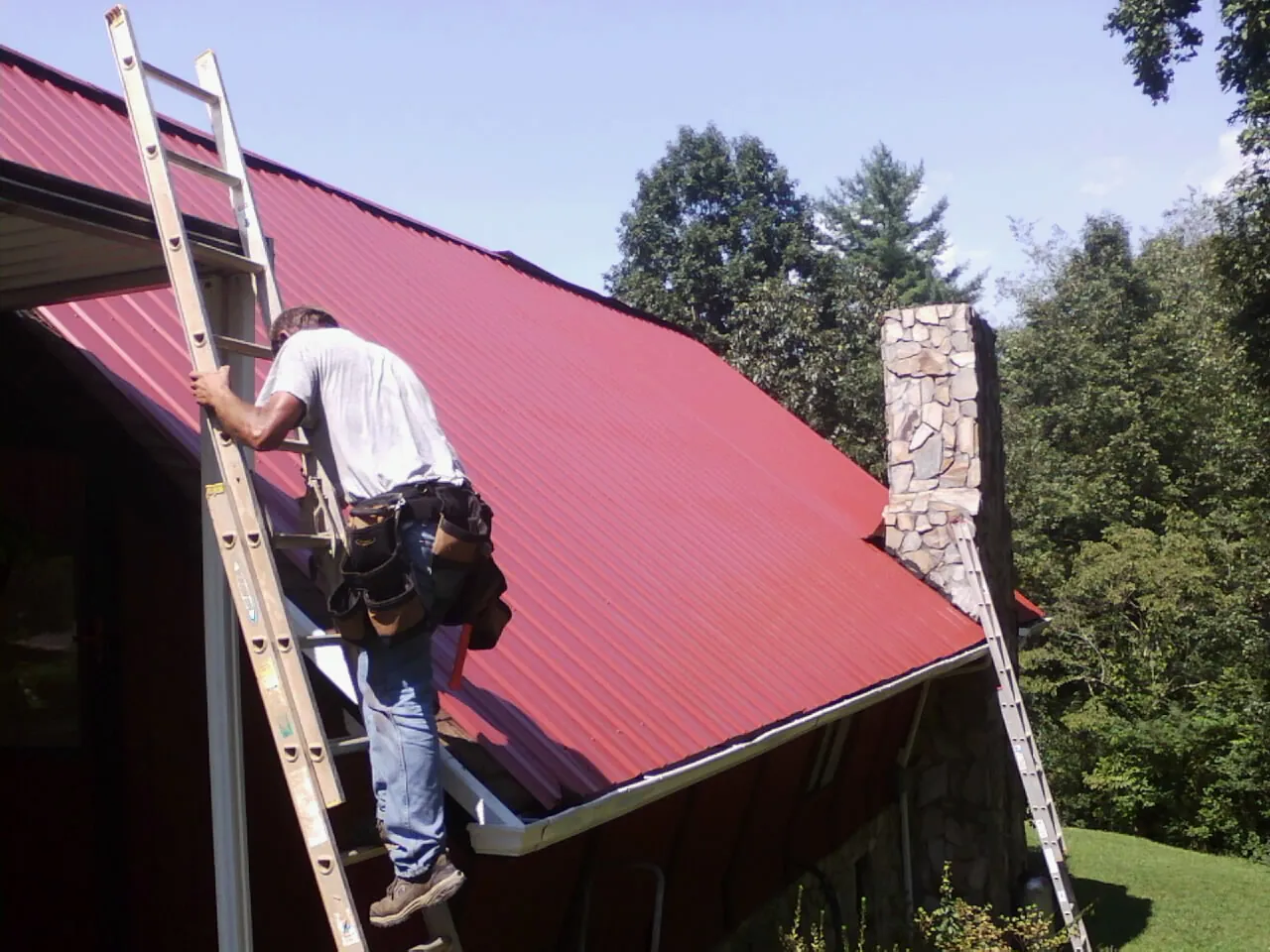 Roofer on a ladder beside a red metal roof and stone chimney — Soffit & Fascia Repair by Roofing Brothers in Western
